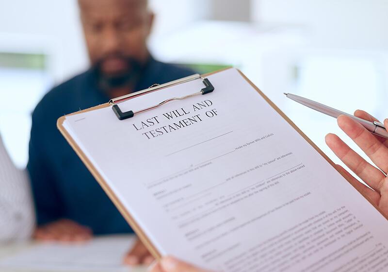 A person holding a "Last Will and Testament" document on a clipboard, with another person out of focus in the background