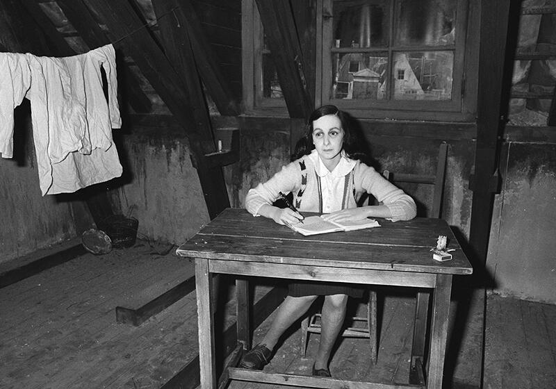 A wax figure of Anne Frank sitting at a desk in a room