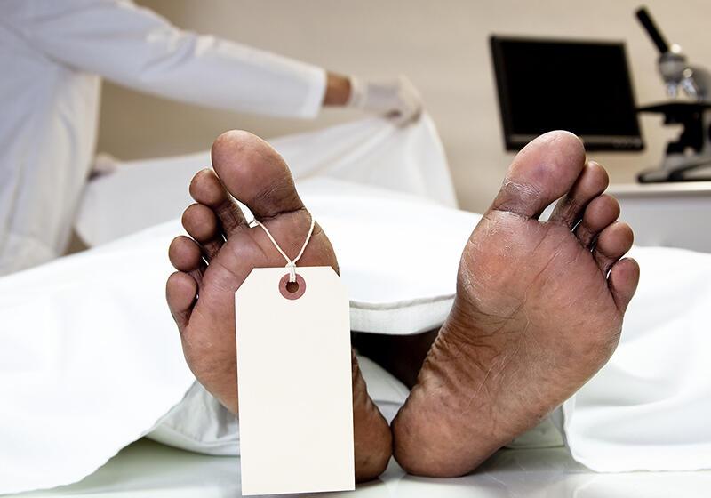 Feet of a deceased person with a toe tag in a morgue, with a person in medical attire in the background