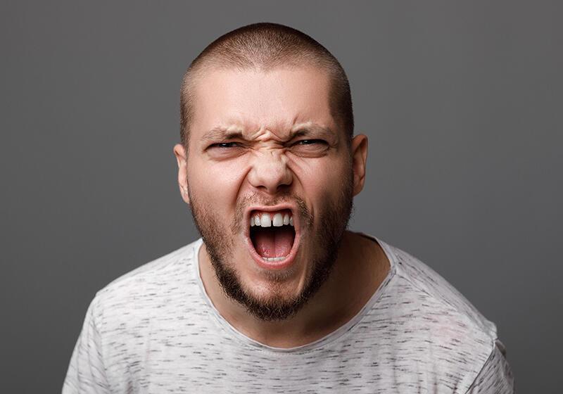 A man with a shaved head and beard screams with an angry expression against a gray background