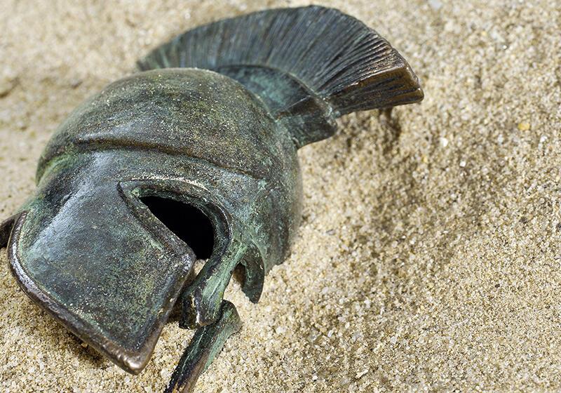 A weathered bronze Greek Corinthian helmet resting in sand