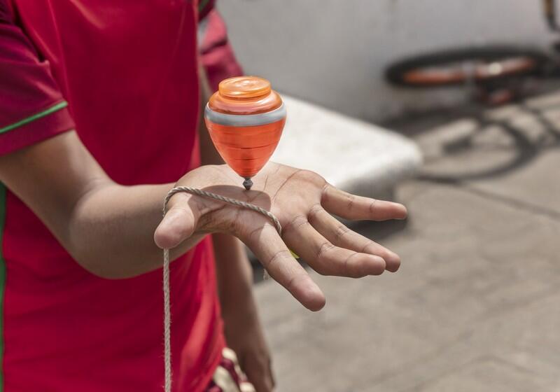 A close-up shot shows a person's open hand balancing a spinning orange and grey plastic top on their palm. The top is supported by a thick string wrapped around the fingers, while the person wears a red shirt with green trim