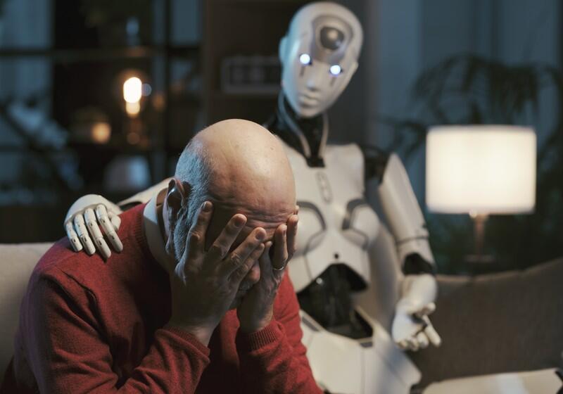 An elderly, bald man sits on a couch covering his face in distress while a humanoid robot places a comforting hand on his shoulder