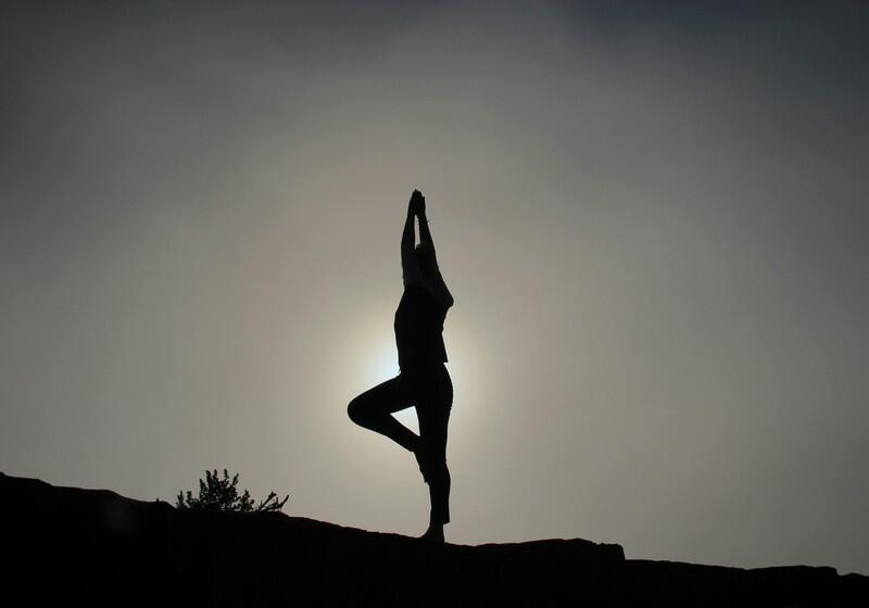 A silhouette of a person performing the yoga tree pose on a rocky hill against a hazy, backlit sky with the sun glowing behind them