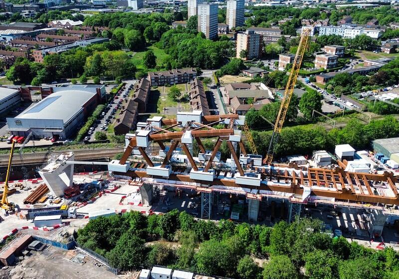 An aerial drone photograph capturing the extensive construction site of a massive steel truss bridge structure, surrounded by cranes, machinery, and urban residential buildings in the background