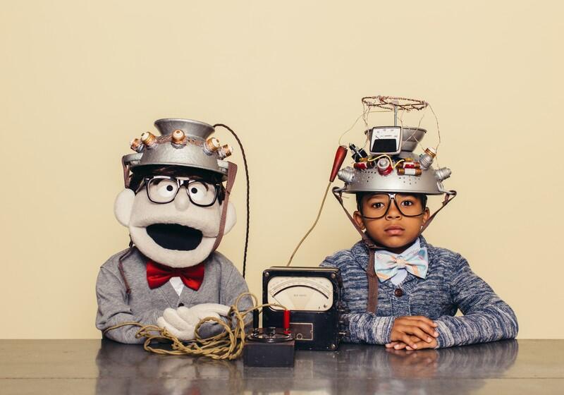 A young boy and a lookalike puppet sit side-by-side at a table wearing matching thick-rimmed glasses, bow ties, and elaborate, wire-covered colanders as "thought-reading" helmets connected to a vintage dial meter