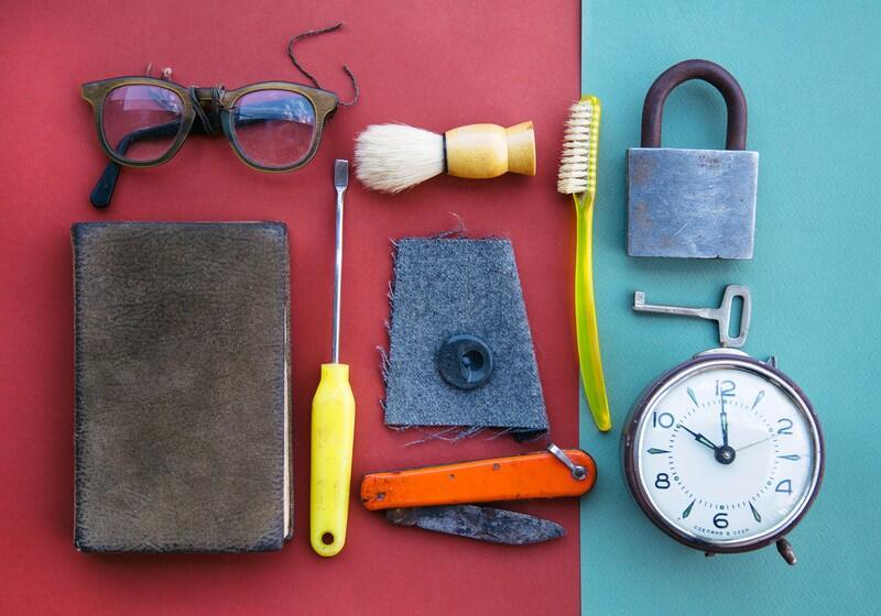 A flat-lay arrangement of vintage items, including a book, eyeglasses, a shaving brush, tools, a padlock with a key, and an alarm clock, neatly organised on a split red and teal background
