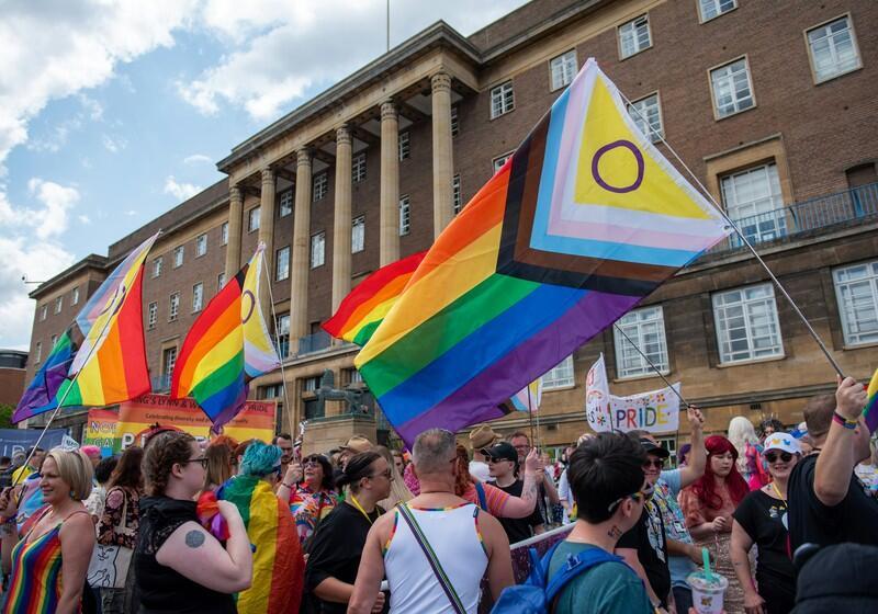 A wide shot captures a lively crowd during a Pride parade gathering outside a large, multi-story brick civic building featuring prominent classical columns