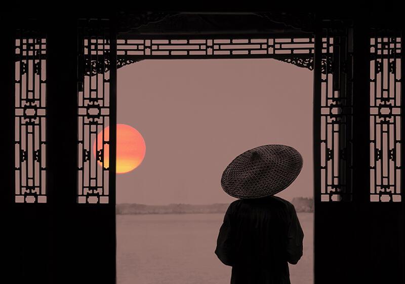 A person wearing a traditional Chinese conical hat stands before an ornate doorway, facing a calm lake at sunset.
