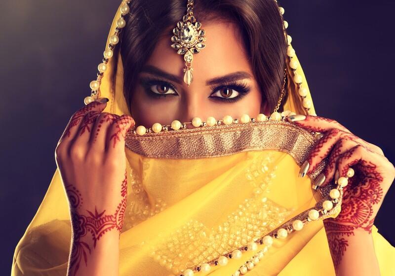 A close-up portrait of a South Asian woman looking directly at the camera, her face partially veiled by a vibrant yellow dupatta adorned with pearls, showcasing intricate henna designs on her hands and a maang tikka on her forehead