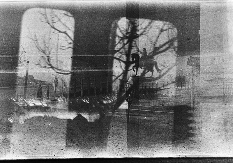 A high-contrast, black-and-white photograph taken through a window, creating a layered, double-exposure effect. Bare tree branches and the silhouette of a person riding a statue of a horse are framed within a window pane