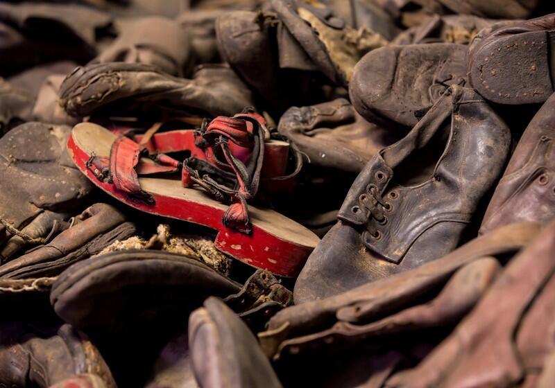 A large, tightly packed pile of old, worn, and dusty leather shoes and sandals belonging to victims of the Holocaust. Amidst the mostly dark brown and black footwear, a single pair of children's sandals with bright red wooden soles and frayed fabric straps stands out prominently near the centre