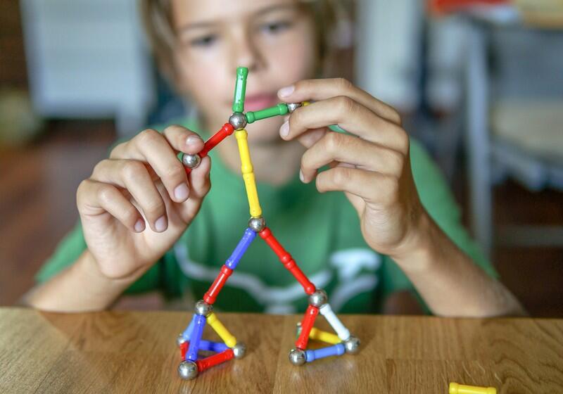 A young boy focuses intently on building a stick figure using a magnetic construction toy set made of colourful rods and metallic spheres on a wooden table