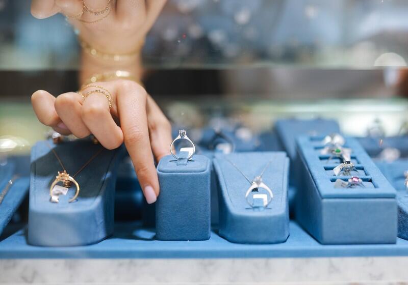 A close-up shot focuses on a person's hand with manicured nails pointing toward a solitaire diamond ring displayed on a blue velvet stand in a jewellery store showcase. The display features various fine jewellery pieces, including necklaces and rings, arranged neatly on matching blue velvet fixtures