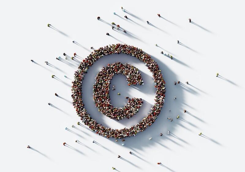 A high-angle, overhead shot shows a large crowd of people gathered on a white surface to form the shape of a copyright symbol