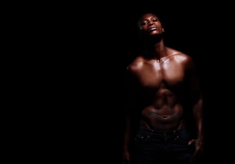 A shirtless, muscular man with dark skin poses against a black background, highlighted by dramatic low-key lighting