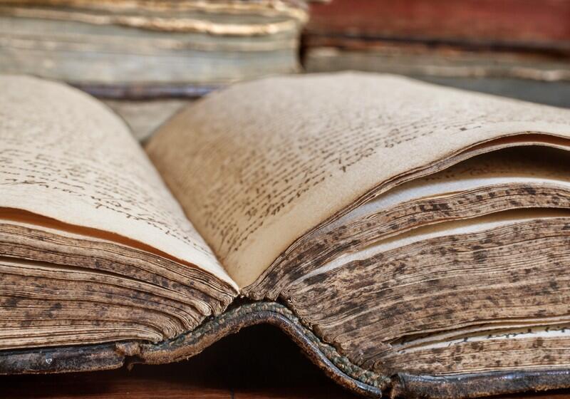 An open, ancient book with thick, weathered pages covered in handwritten script is shown in a close-up view