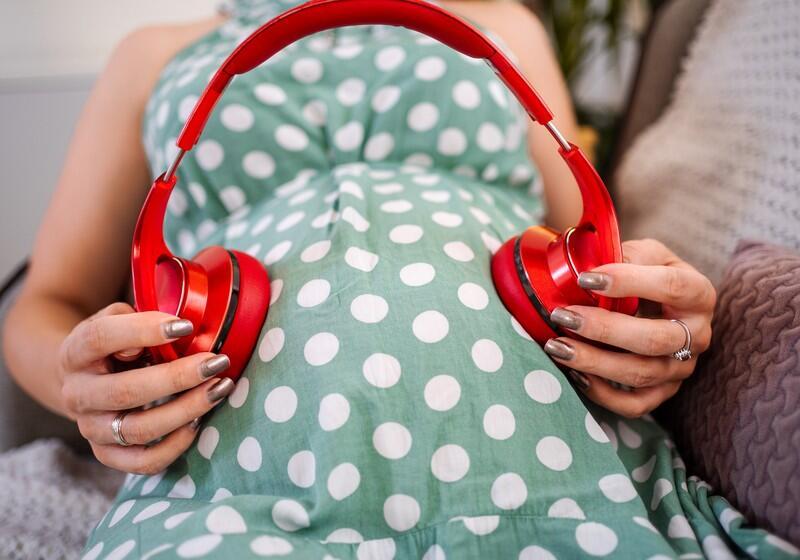 A close-up shot of a pregnant woman wearing a green and white polka dot dress, holding a pair of large, bright red over-ear headphones against her pregnant belly, positioning an ear cup on each side of the bump