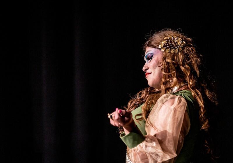 A profile shot of a performer with curly light brown hair, heavy stage makeup, and an ornate gold hair clip smiling while gesturing on a dark stage