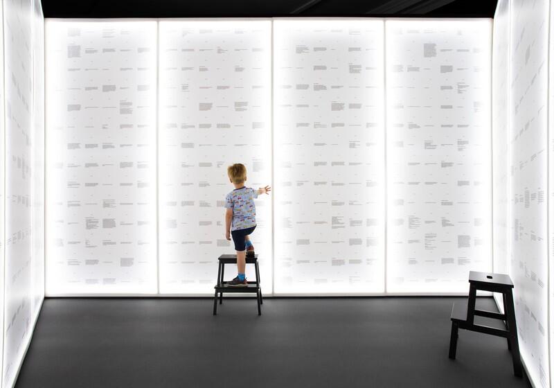 A young child stands on a black step stool with their back to the camera, reaching out to touch one of several large, floor-to-ceiling glowing white panels that form the walls of a room