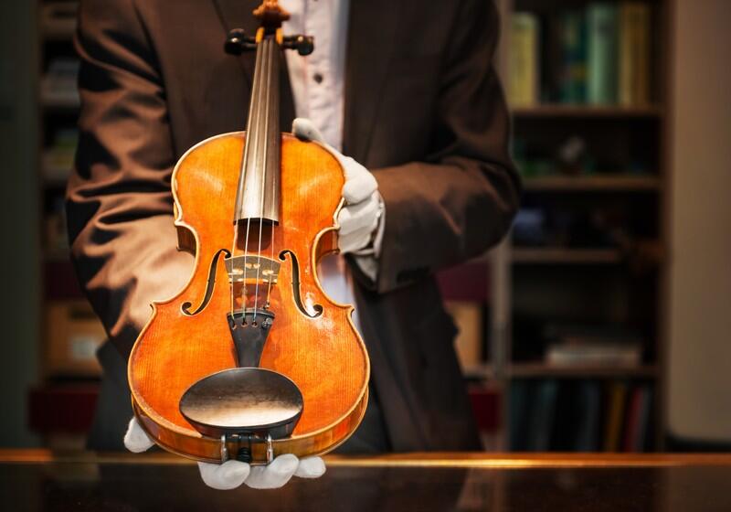 A close-up shot of a person in a brown suit jacket and white gloves holding out a polished wooden violin for display, against a blurred background of a wooden bookshelf filled with books