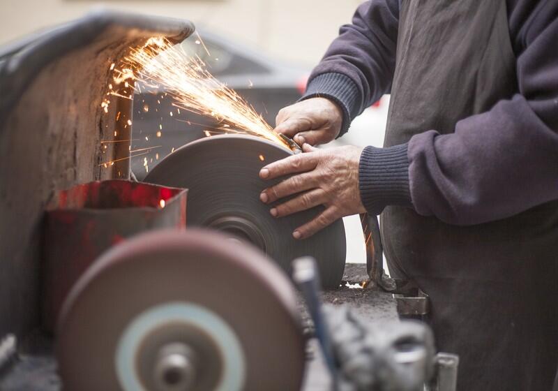 An artisan grinds a metal tool against a large, rotating bench grinder wheel, producing a bright shower of orange sparks. The worker, dressed in a dark apron and long sleeves, carefully guides the metal with bare hands. In the foreground, another grinding wheel sits out of focus on the workbench
