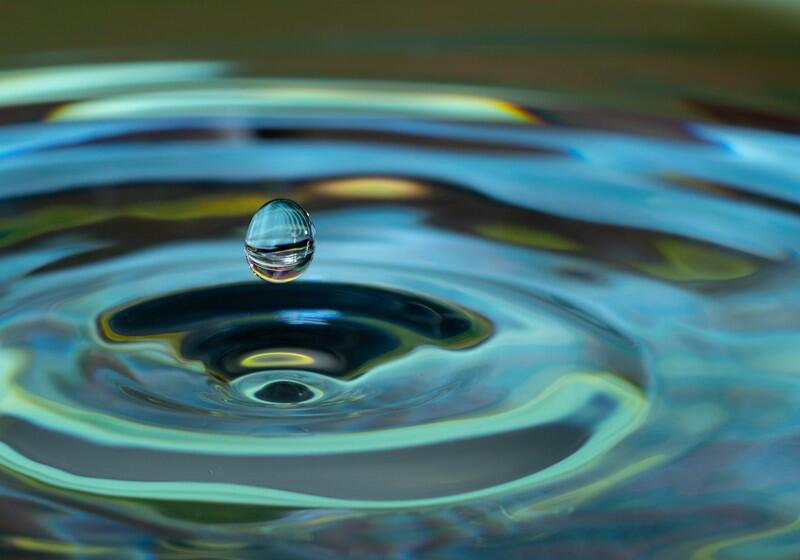 A close-up photograph captures a single water droplet suspended in the air just above a rippling water surface. The droplet reflects its colourful surroundings. Below it, concentric, circular ripples expand across the fluid surface, creating waves of light teal, deep blue, and pale yellow-green. The background shows a soft, out-of-focus blur of the same fluid colours