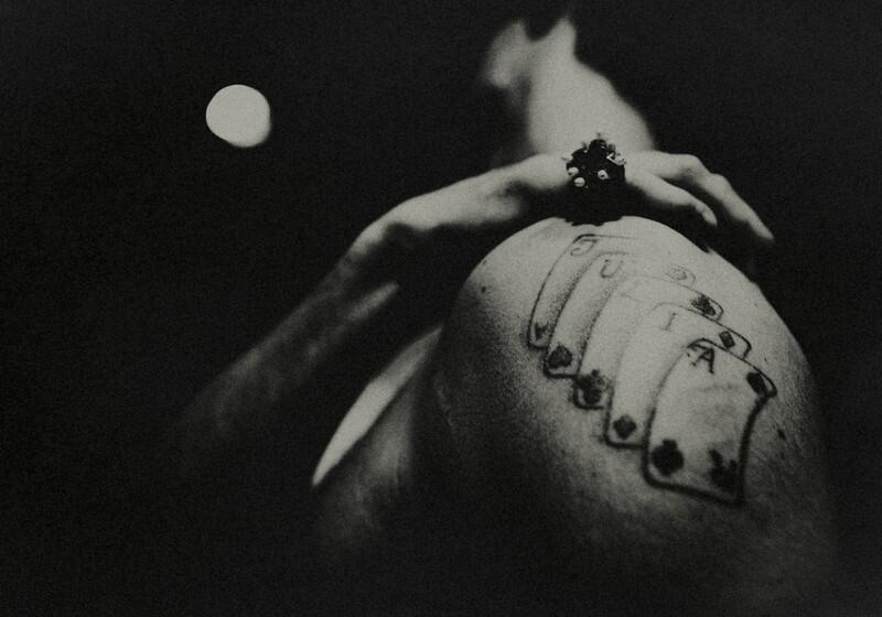 A high-contrast, grainy black and white photograph captures a person from a low angle, emphasising a tattoo on their upper arm and shoulder