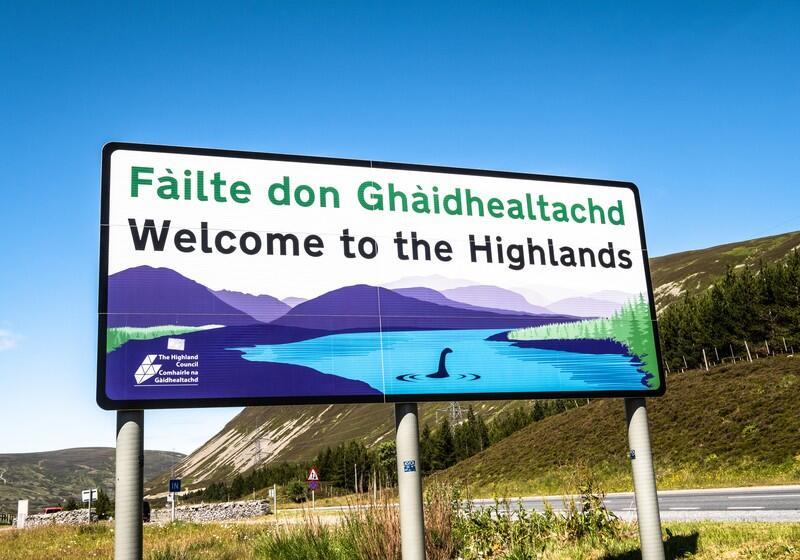 A large rectangular roadside welcome sign stands against a bright, clear blue sky, reading "Fàilte don Ghàidhealachd" in green text and "Welcome to the Highlands" in bold black text below it. The sign features an illustration of a scenic Scottish landscape with purple mountains, a blue lake, green pine trees, and a silhouette resembling the Loch Ness Monster