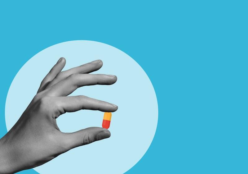 A stylised illustration features a black-and-white hand holding a small capsule pill against a bright blue background. The capsule is coloured with an orange top and a red bottom. A light blue circle serves as a backdrop behind the hand, highlighting the gesture