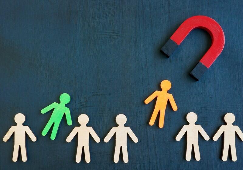 A top-down view of several small, wooden human-shaped cutouts arranged against a dark, textured blue background. A large red and black horseshoe magnet in the upper right corner appears to draw an orange figure away from a group of plain beige figures. To the left, a bright green figure stands out, tilted away from the others