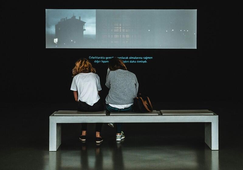 Two people sit on a bench from behind, watching a video projection with subtitles in a dark gallery space