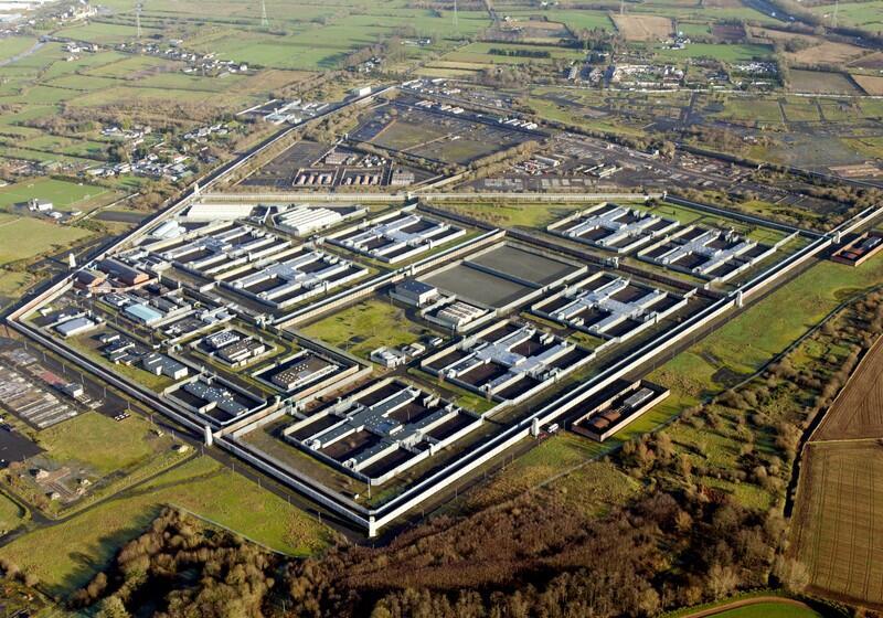 An aerial view of the HM Prison Maze complex, featuring its distinctive H-block layout with several H-shaped prison buildings surrounded by high security walls, watchtowers, and open green fields