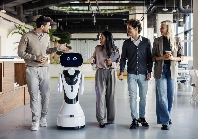 A small, futuristic white robot with digital blue eyes walks through a modern, open-plan office alongside four colleagues who are smiling and conversing