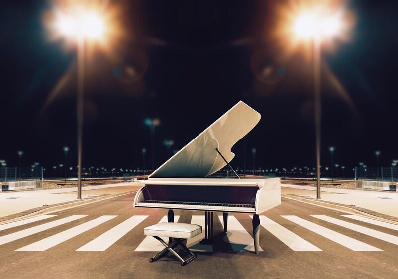 A white grand piano with its lid open sits in the middle of a pedestrian crosswalk on an empty city street at night, accompanied by a matching white piano bench. Two bright streetlights illuminate the scene from both sides, creating a dramatic, symmetrical glow against the dark sky