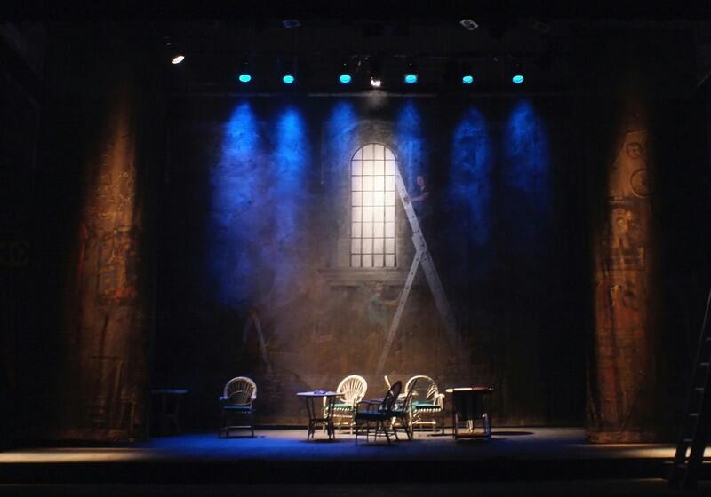 A theatre stage set designed to look like a stone wall courtyard or interior, illuminated by blue spotlights from above. In the centre backdrop, a painted arched window is lit with white light, with a ladder leaning against it. On the stage floor below, several wicker chairs and small round tables are arranged. Two large, dark columns frame the left and right sides of the stage