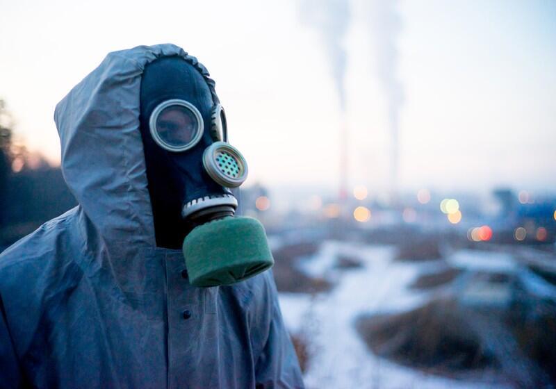 A person wearing a hooded protective jacket and a vintage gas mask stands outdoors against a blurred industrial background. The black rubber mask covers their entire face, featuring circular eye lenses, a perforated metal speech diaphragm, and a green cylindrical filter attached to the bottom. In the soft-focus distance, smoke plumes rise from industrial chimneys under a pale sky