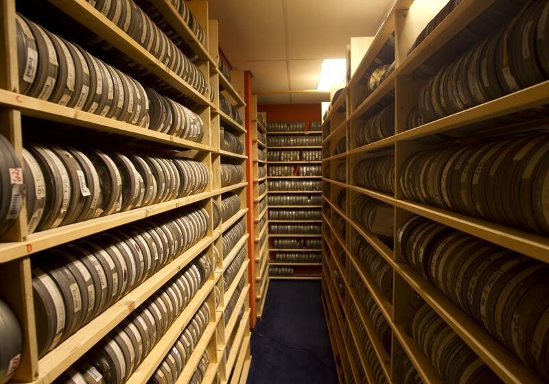 Two long rows of tall wooden shelves filled with rows of film canisters pack a narrow storage room with a blue carpeted floor. The shelves on the left and right extend forward into the foreground, while another shelf stands against the back wall