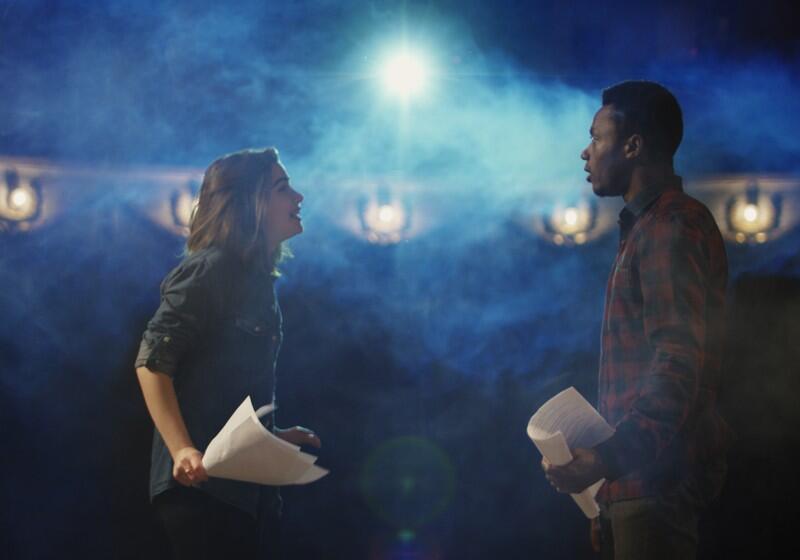 A young woman and man rehearse with scripts on a dimly lit theatre stage, illuminated by a bright spotlight amidst blue stage smoke
