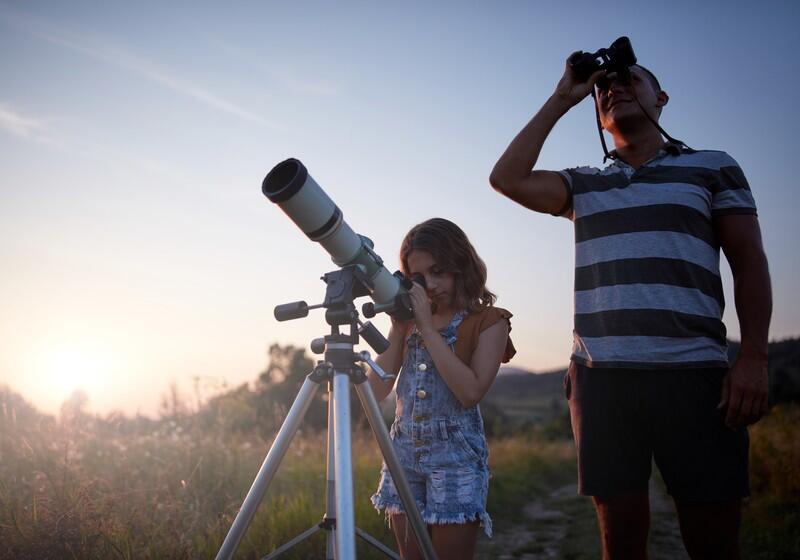 A father and his young daughter stargazing together in an open grassy field during sunset. The daughter looks through a white telescope mounted on a tripod, while her father stands beside her, looking up at the sky through a pair of binoculars