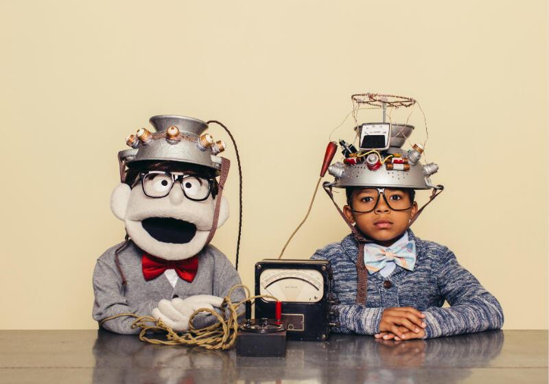 Two children, one real and one a puppet, wearing homemade metal helmets with wires sit at a table, connected to a small device.