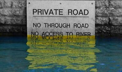 A "Private Road" sign, partially submerged in water, reads "NO THROUGH ROAD NO ACCESS TO RIVER"