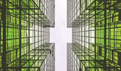 Looking up at the modern green glass facade of tall buildings meeting in a cross shape against a bright sky