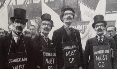 Participants at a march wearing moustachioed masks and signs saying 'Chamberlain must go'