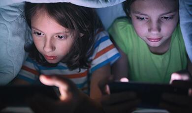 Two children on digital devices under bed covers