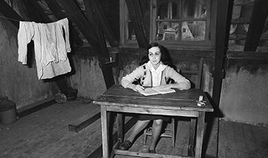 A wax figure of Anne Frank sitting at a desk in a room