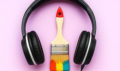 Black over-ear headphones with a paint brush featuring a red, yellow, and blue tip positioned beneath them on a pink background