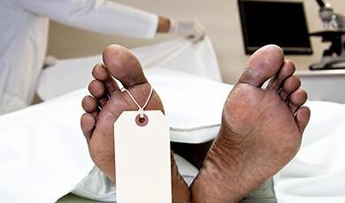 Feet of a deceased person with a toe tag in a morgue, with a person in medical attire in the background
