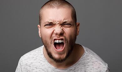 A man with a shaved head and beard screams with an angry expression against a gray background
