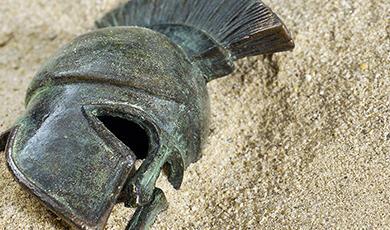 A weathered bronze Greek Corinthian helmet resting in sand